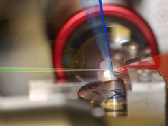 A laser pulse (red) hits helium atoms (blue), streaming from a nozzle with supersonic velocity. A very compact and controlled difference in density (dark blue ray) develops due to the partial obscuring of the nozzle by a razor blade. Precisely at this difference in density, the laser pulse hits the helium atoms, separates the electrons and accelerates them to nearly the speed of light. Since the electrons are all separated at the same location and same time from the atoms, they nearly gain the same energy.