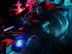 Photo showing the vacuum chamber used to measure the 2S-4P transition frequency in atomic hydrogen. The purple glow in the back stems from the microwave discharge that dissociates hydrogen molecules into hydrogen atoms. The blue light in the front is fluorescence of the vacuum viewport from the ultraviolet laser that excites the atoms to the 2S state. The turquoise blue glow is stray light from the laser system used to measure the frequency of the 2S-4P transition. (Credit: Axel Beyer / MPQ)
